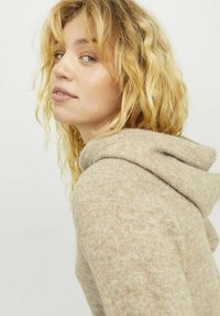 Felpa beige lavorata a maglia con una texture morbida, caratterizzata da un collo ampio e una vestibilità rilassata. Capelli ricci incorniciano il viso, evidenziando lo stile informale del capo.