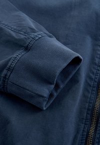 Manche de veste bleu marine avec poignet côtelé à côté d'une fermeture éclair métallique visible sur un fond en tissu lisse.