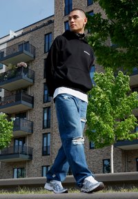 Sweat à capuche noir, T-shirt gris clair en dessous, jean bleu déchiré et chaussures de sport blanches avec des accents gris. Cadre urbain.