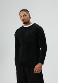 Zwarte kabelgebreide trui met een getextureerde afwerking, ronde hals en lange mouwen, gecombineerd met loszittende zwarte broek.