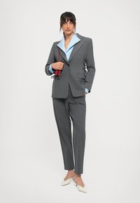 Woman in gray suit with blue shirt, holding small red purse, wearing white heeled shoes, standing against white background.