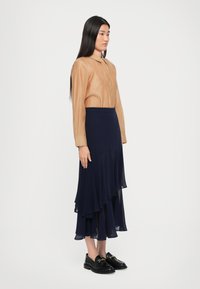 Femme aux longs cheveux noirs portant un chemisier beige à boutons, une jupe midi superposée marine et des mocassins noirs, se tenant devant un mur blanc uni.