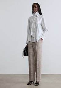 Shiny silver blouse with a high collar and bow detail, paired with patterned beige and pink wide-leg trousers; black handbag and sandals.