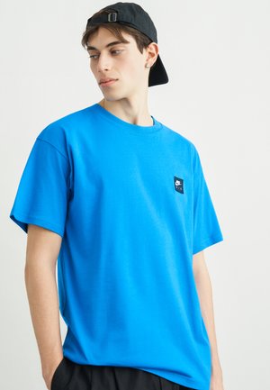 Joven con una camiseta Nike Air azul brillante y una gorra negra al revés, mirando hacia un lado con las manos en los bolsillos.