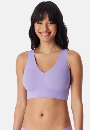 Glimlachende vrouw met lang donker haar die een lichtpaars mouwloos cropped top draagt tegen een effen witte achtergrond.