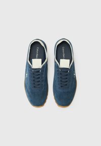 Blue suede sneakers with rounded toe, laced design, white inner lining, and tan rubber outsole. Features logo on the side.