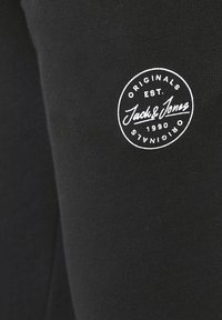 Schwarze Jogginghose mit einer weichen Textur, die ein rundes weißes Logo mit Text und Gründungsjahr auf dem linken Bein zeigt.