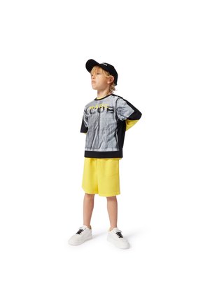 Grafisk t-skjorte med fargene svart, grå og gul, kombinert med lyse gule shorts og hvite joggesko. Akkompagneres av en svart caps.