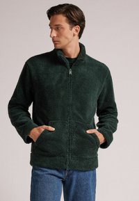 Zielona polarowa kurtka na zamek z wysokim kołnierzem, dłoniami i dwiema przednimi kieszeniami; miękki materiał i swobodny krój.