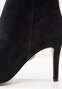 Bottine en daim noir avec un talon haut fin et un design lisse et sans couture visible de profil.