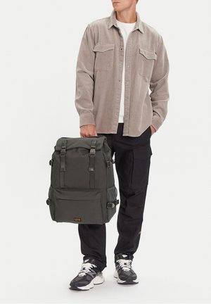 Mann trägt beige Cordhemd und schwarze Cargohosen, hält schwarzen Rucksack seitlich, steht vor weißem Hintergrund.