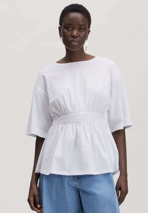 Dark-skinned person wearing a white gathered waist short-sleeve top and light blue jeans, standing against a plain background.