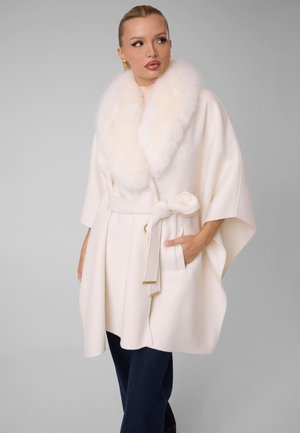 Femme portant un manteau blanc ceinturé avec un grand col duveteux, une main dans la poche, regardant sur le côté sur un fond gris uni.