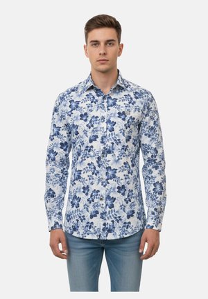 Joven con camisa blanca de botones con patrones florales azules y vaqueros azul claro, de pie frente a un fondo blanco liso.