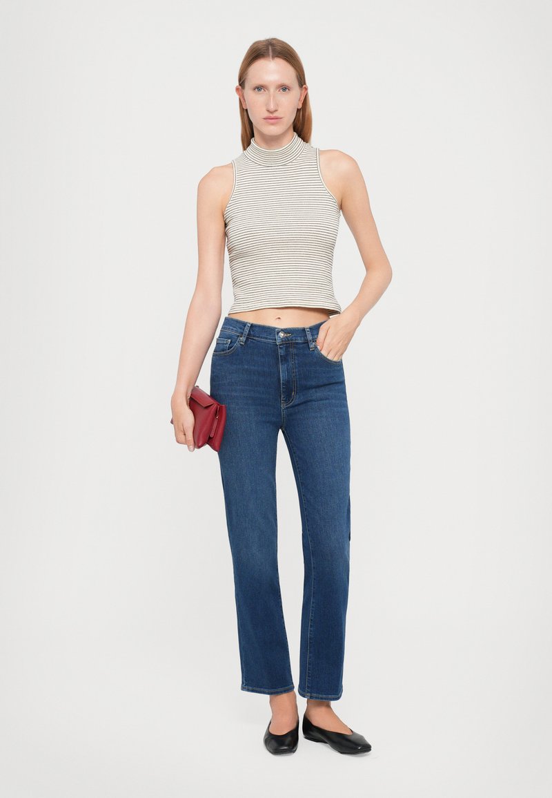 Gestreepte, mouwloze top met hoge hals, gecombineerd met donkerblauwe rechte jeans. Het model houdt een kleine rode clutch vast en draagt zwarte ballerina's.