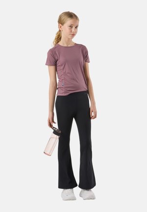 Ung pige iført en støvet lilla kortærmet top, sorte udvidede bukser, hvide sneakers, holder en gennemsigtig vandflaske med sort låg.