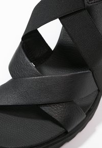 Sandale noire avec des sangles croisées en cuir lisse et matériau élastique, dotée d'une semelle intérieure texturée et d'une semelle plate.