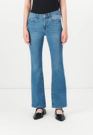 Moteris vilki šviesiai mėlynas plataus kirpimo džinsus, juodą be rankovių palaidinę ir juodus batelius su kvadratine nosimi ant vientisos baltos fono.