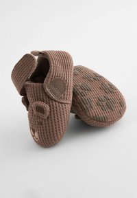 Papuci pentru bebeluși dintr-un material fabricat maro, cu o țesătură texturată de tip waffle, având o curea cu Velcro și detalii în formă de labă de animal pe talpă.