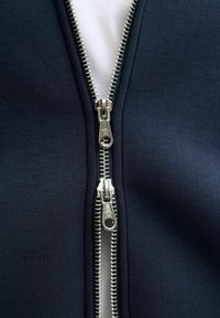 Marinblå zip-up jacka med dubbla silvriga dragkedjor, med en slät tygtextur och kontrasterande vitt foder vid öppningen.