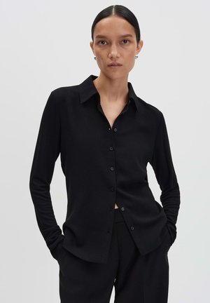 Vrouw met donker haar in een lage bun, gekleed in een zwarte blouse met knopen en zwarte broek, staand met handen in de zakken tegen een witte achtergrond.