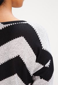 Person, die einen schwarz-grauen Pullover mit Zickzackmuster trägt, von hinten und über die Schulter gegen einen schlichten hellen Hintergrund gezeigt.