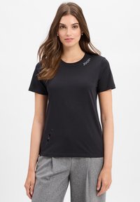 Zwarte katoenen T-shirt met een ronde halslijn, korte mouwen, een gestikt logo en een klein snitdetail aan de zoom. Gecombineerd met lichtgrijze broeken.