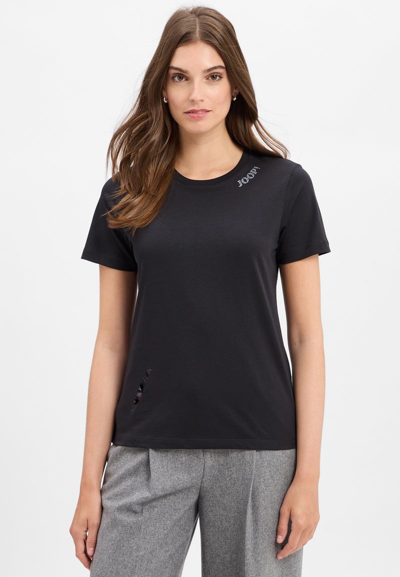 Zwarte katoenen T-shirt met een ronde halslijn, korte mouwen, een gestikt logo en een klein snitdetail aan de zoom. Gecombineerd met lichtgrijze broeken.