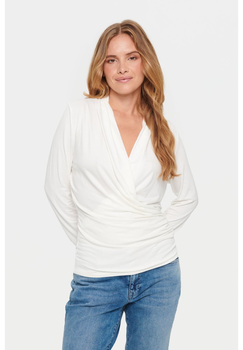 White wrap top with long sleeves, deep V-neck, soft texture, and gathered side detail, paired with blue denim jeans.