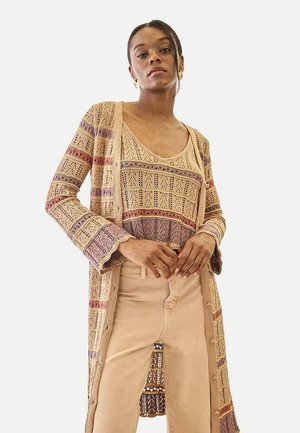 Mujer vestida con un cárdigan de punto beige con estampado morado y una blusa sin mangas a juego, combinados con pantalones beige de tiro alto, de pie frente a un fondo blanco.