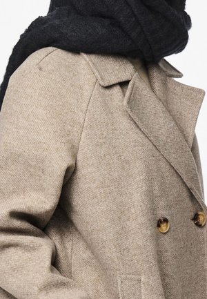 Gros plan sur un manteau en laine beige avec un col cranté, des boutons croisés et une écharpe noire texturée enroulée autour du cou.