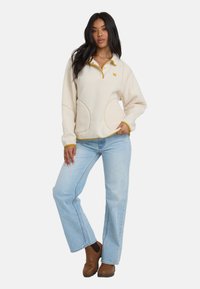 Frau trägt einen cremefarbenen Fleece-Pullover mit gelbem Saum, hellblaue Jeans und braune Stiefeletten. Sie steht mit einer Hand in der Tasche.