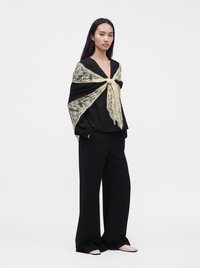 Femme debout portant un costume noir avec un pantalon à jambes larges et un châle en dentelle noué en nœud sur les épaules sur fond blanc.