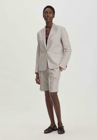 Lichtbeige linnen blazer met een enkele knop, gecombineerd met bijpassende shorts. Het model draagt een zwarte top en zwarte platte sandalen. Eenvoudig ontwerp.