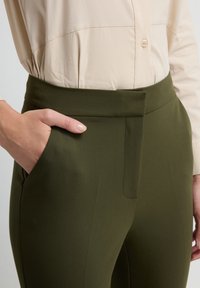 Schlanke, olivgrüne Hose mit flachem Bund, ausgestattet mit einer Seitentasche und glattem Stoff. Kombiniert mit einer hellbeigen Bluse.