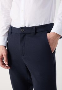 Homme portant un pantalon habillé bleu marine avec une ceinture boutonnée et une chemise blanche à manches longues, main droite dans la poche, main gauche détendue le long du corps.