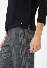 Zwarte lange mouw top met een zijsplit en een zilveren logo-accent nabij de zoom, gecombineerd met grijs geruite gepaste broek met zakken.