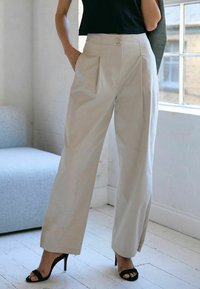 Un pantalon beige à jambes larges avec des plis devant et deux fermetures à bouton, fabriqué dans un tissu lisse. Assorti à des sandales à talons hauts noires.