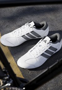 Bílé tenisky Adidas s černými pruhy a tkaničkami, umístěné na asfaltu blízko žluté čáry, částečně rámované černým kolem.
