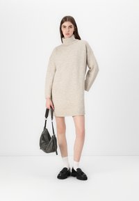 Beige høyhalset kjole med lange ermer, rett snitt, kombinert med en grå skulderveske og svarte loafers, mot en hvit bakgrunn.