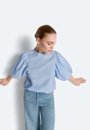 Kobieta o jasnobrązowych włosach związanych w kok, ubrana w jasnoniebieską bluzkę z bufkami i dżinsy, opierająca się plecami z rękami o białą ścianę, patrząca w dół w prawo.