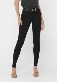 Vaqueros skinny negros con una textura suave, cintura alta, bolsillos frontales y un cinturón. Combinados con sandalias deslizables en color tostado.