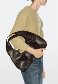 Frau im beigen Pullover und blauer Jeans, die eine große, dunkelbraune Patchwork-Ledertasche unter dem Arm hält, Seitenansicht.
