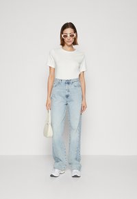 Weißes kurzärmeliges T-Shirt, kombiniert mit hellblauen hochgeschnittenen Jeans. Dazu weiße Sonnenbrille und eine kleine cremefarbene Tasche.