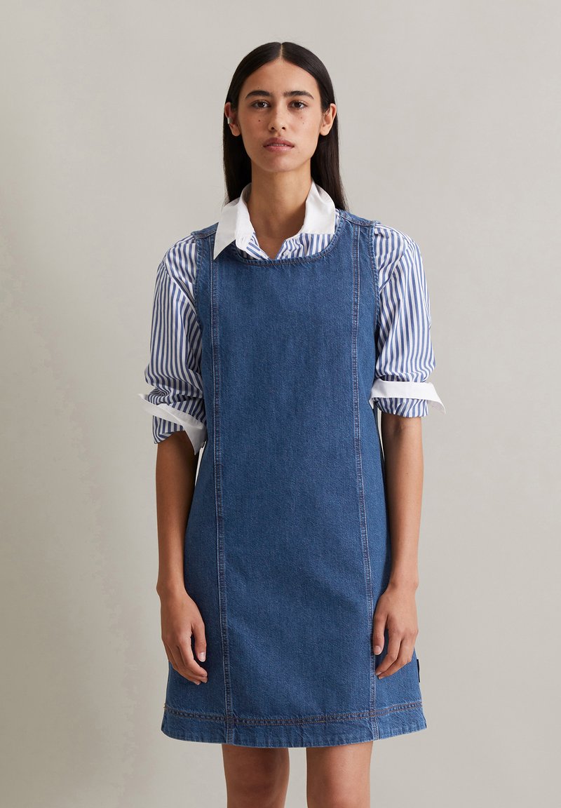 Denim jurk in een rechte snit met een ronde halslijn en decoratieve stiksels, gedragen over een blauw-wit gestreept shirt.