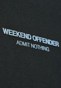Tessuto nero con testo bianco in rilievo che dice "WEEKEND OFFENDER" sopra il testo più piccolo "ADMIT NOTHING".