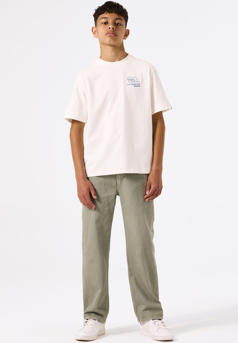 Crème katoenen T-shirt met korte mouwen en een kleine blauwe grafische afbeelding op de borst, gecombineerd met lichtgroene broek met rechte pijpen en witte sneakers.