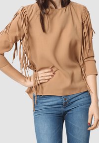 Mujer con blusa de mangas con flecos en tono marrón y vaqueros azules con cierre de botones, con una mano en la cadera y uñas pintadas de un rojo oscuro.