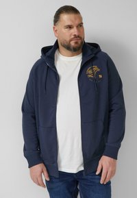 Marineblauwe hoodie met ritssluiting, een voorzak en een capuchon met trekkoord. Heeft een goudkleurig logo op de linkerborst. Gedragen over een wit T-shirt.