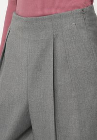 Grå skreddersydde bukser med folder og glidelås på siden, laget av et lett materiale, som viser en glatt tekstur og en strukturert design.